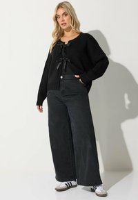 Svart stickad cardigan med rund halsringning, prydd med satinband och vida ärmar, i kombination med lösa svarta denimjeans och sneakers.