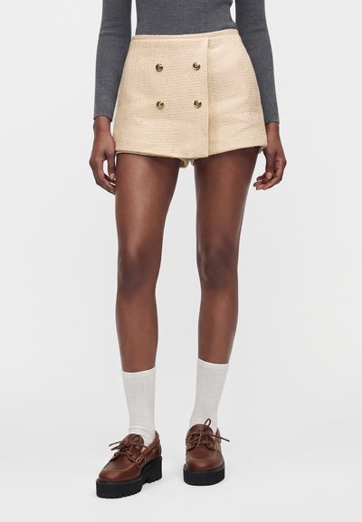 Femme portant un short beige texturé avec des boutons dorés, un haut gris à manches longues, des chaussettes blanches et des chaussures à plateforme marron, debout.