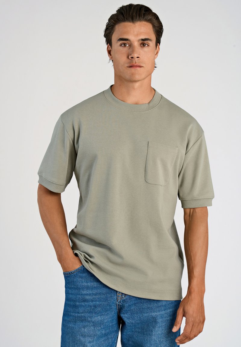 Ljust grå kortärmad t-shirt med rund nacke och en framficka, tillsammans med blå denimjeans. Slät tygkänsla.