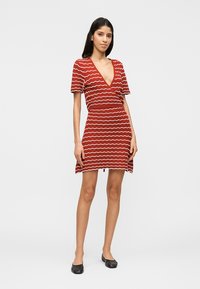 Frau steht und trägt ein rotes Wickelkleid mit kurzen Ärmeln, weißen wellenförmigen Streifen und schwarzen flachen Schuhen vor einem schlichten hellen Hintergrund.