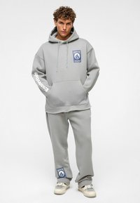 Grijze hoodie en sweatpants van zachte stof. De hoodie heeft een voorzak en een grafische patch; de broek heeft zijzakken en een boord aan de onderkant.