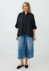 Black oversized shirt with a collar and rolled sleeves, paired with wide-leg blue denim jeans featuring a frayed hem. Black slip-on shoes.