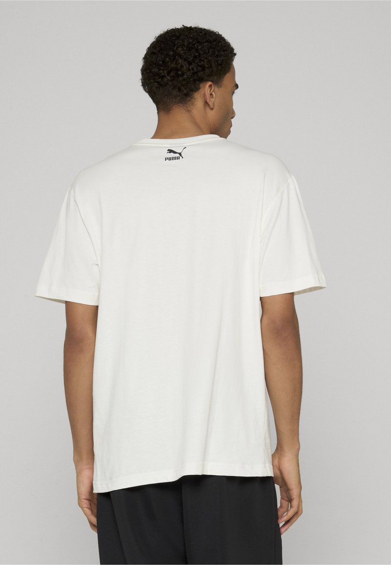 Muž otočený chrbtom, oblečený v voľnom bielom tričku Puma s logom umiestneným v strede pri límci a čiernych nohaviciach na jednoduchej pozadí.