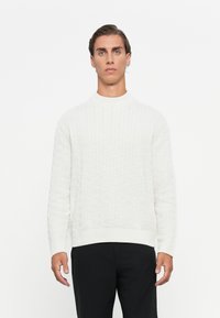 Modello maschile con capelli all'indietro, vestito con un maglione bianco a trecce e pantaloni neri, su uno sfondo bianco uniforme.