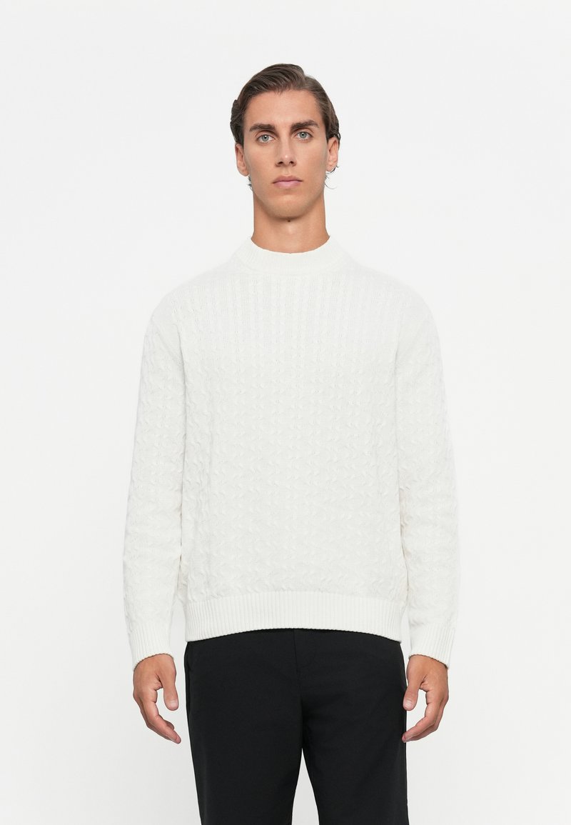 Modello maschile con capelli all'indietro, vestito con un maglione bianco a trecce e pantaloni neri, su uno sfondo bianco uniforme.