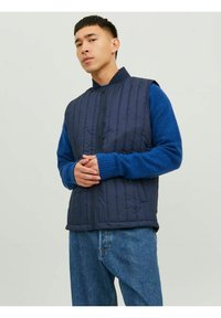 Mornarsko quilted telovnik z visokim ovratnikom, v kombinaciji z modrim svetrom z dolgimi rokavi in sproščenimi jeanskimi hlačami; stoječ položaj.