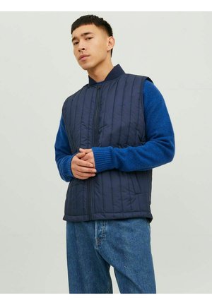 Marineblauwe gevoerde vest met hoge kraag, gedragen met een blauwe lange mouwen trui en een relaxed-fit spijkerbroek; staande pose.