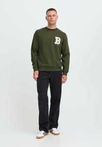 Mann som står, iført grønn genser med bokstaven B, svarte jeans og hvite joggesko mot en ensfarget lys bakgrunn.