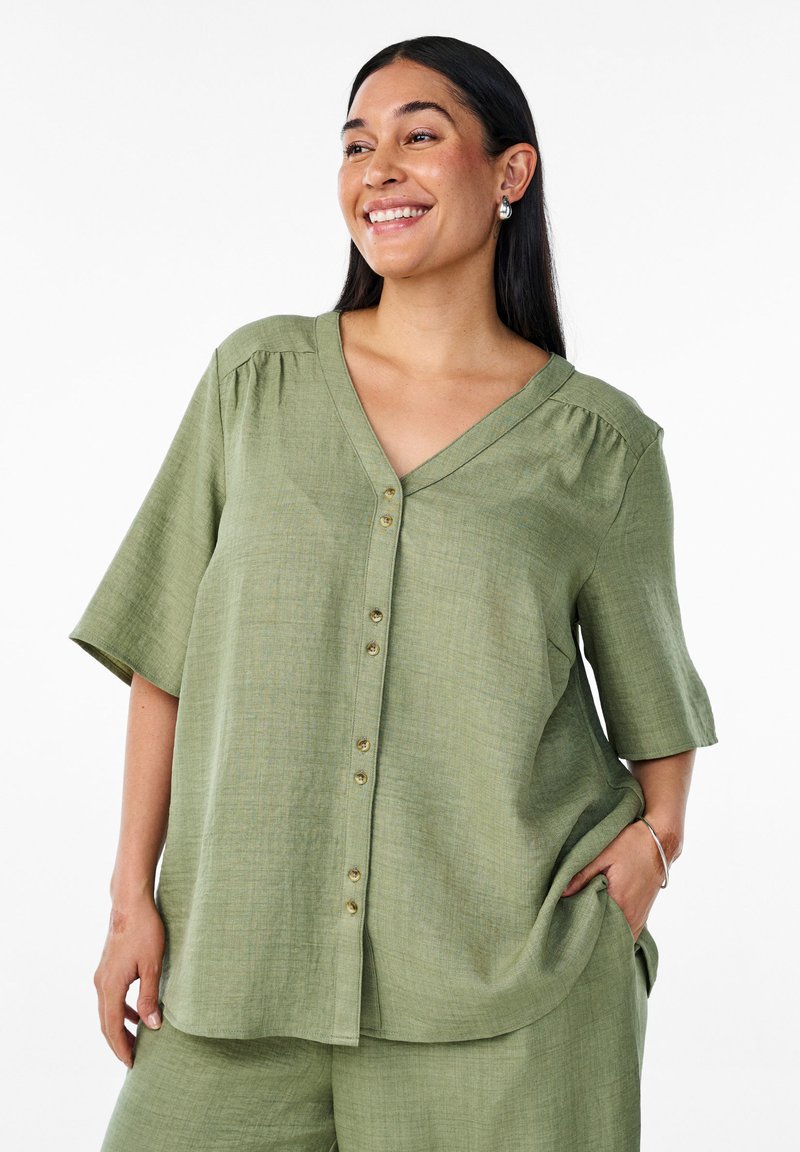 Camicia verde chiaro a maniche corte con scollo a V, chiusura a bottoni e tessuto strutturato. Presenta una vestibilità rilassata e leggere arricciature sulle spalle.