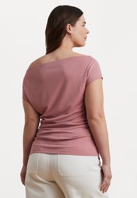 Roza off-shoulder majica iz mehkega materiala s kratkimi rokavi in oprijetim krojem, v kombinaciji z belimi hlačami. Teksture in drapirane oblikovne podrobnosti.