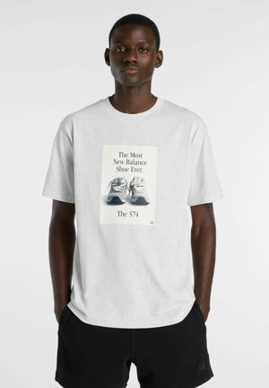 Mann trägt hellgraues T-Shirt mit New Balance 574 Schuh-Grafik und schwarze Shorts, steht vor weißem Hintergrund.