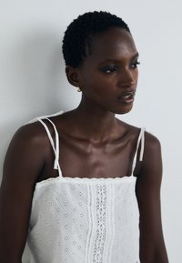 Top scurt alb din material cu perforații, cu bretele subțiri, având modele florale intricate și un material texturat. Pielea fină și bijuteriile subtile completează aspectul.