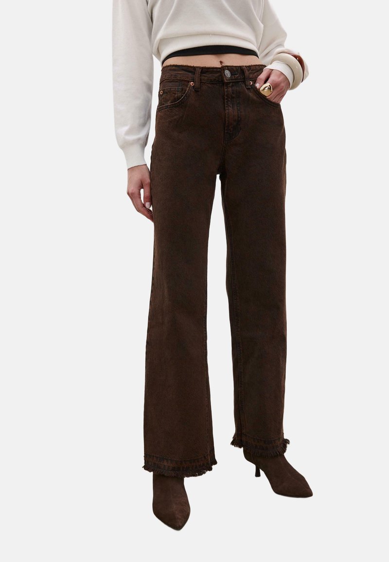 Femme portant un jean large marron foncé aux ourlets effilochés, un pull court blanc, des bottines marron à bouts pointus et des bracelets épais.