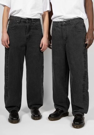 Jeans negros de talle alto y pierna ancha, con pliegues, que presentan un estampado de texto lateral y bolsillos traseros, combinados con zapatos de cuero negros.