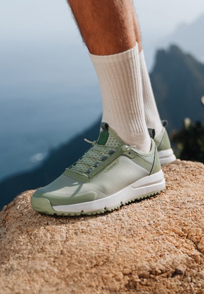 Ljusgröna sportskor med nät- och syntetmaterial, med vit sula och snören, placerade på en stenig yta mot en suddig bakgrund.