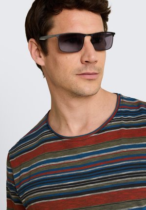 Mężczyzna noszący ciemne prostokątne okulary przeciwsłoneczne i wielokolorową, paskowaną koszulkę z okrągłym dekoltem, spogląda lekko na bok.