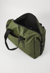 Sac de sport vert olive avec des accents noirs. Dispose d'une ouverture zippée, d'une poignée courte et d'une poche latérale pour le rangement. Texture lisse.