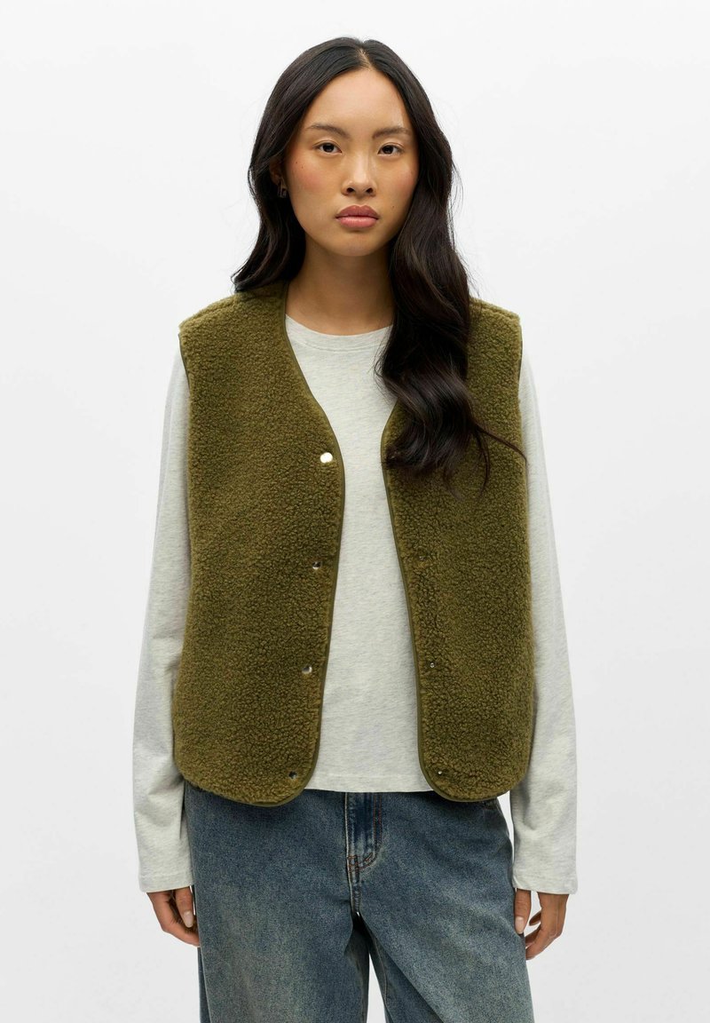 Femme aux longs cheveux foncés porte un gilet en polaire vert olive par-dessus une chemise blanche à manches longues et un jean bleu ample sur un fond blanc uni.
