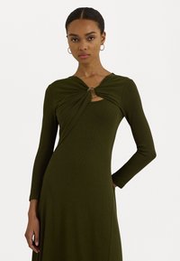 Kvinna iklädd en olivgrön långärmad klänning med vriden halsringning och metallringdetalj, stående med ena handen på höften mot en vit bakgrund.