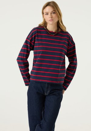 Frau mit langen blonden Haaren, die ein marineblau-rot gestreiftes Langarmshirt, dunkle Jeans und eine auffällige Perlenkette trägt, steht entspannt.