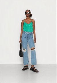Camisola de alças com padrão floral verde, jeans rasgados de cintura alta em azul claro, sandálias de plataforma pretas e uma bolsa de ombro preta.