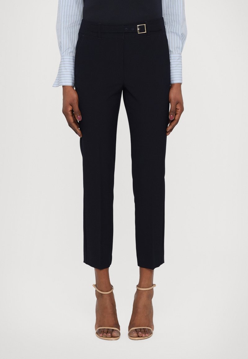 Femme portant un pantalon noir tailleur longueur cheville avec une fine ceinture, une chemise rayée bleu clair et des talons ouverts beige sur un fond uni.