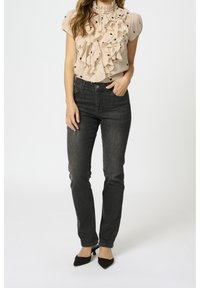 Beige blouse met ruffles en zwarte stippen, ingestopt in donkergrijze jeans. Model draagt zwarte puntige hakken, met haar handen op één heup.