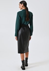 Donna con capelli lunghi e scuri, indossa una blusa verde, una gonna a matita in pelle nera e stivali neri, in piedi e di spalle su uno sfondo neutro.
