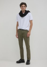 T-shirt bianco a maniche corte, pantaloni aderenti verde oliva, sciarpa nera in maglia appoggiata intorno al collo, scarpe nere stringate. Design semplice e moderno.