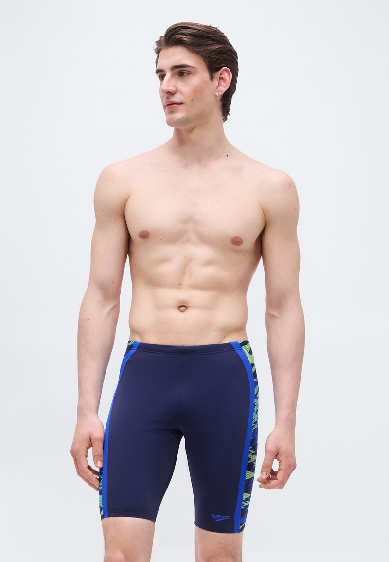 Άνδρας χωρίς πουκάμισο που φοράει μπλε σκούρα μαγιό τύπου jammer με πλαϊνές πλευρές σε μπλε και πράσινο μοτίβο, στέκεται μπροστά σε ένα απλό ανοιχτόχρωμο φόντο.