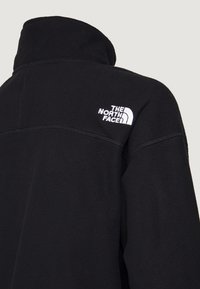 Chaqueta de forro polar negra con cuello alto, que presenta un logo bordado en blanco en el hombro izquierdo y detalles de costura en la parte posterior.