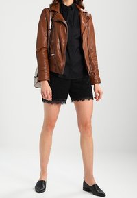 Giubbotto in pelle marrone sopra una maglietta nera, abbinato a pantaloni corti neri con pizzo e mocassini a punta neri, su uno sfondo neutro.