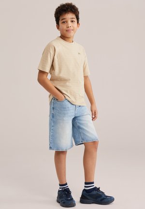 Niño de pie con las manos en los bolsillos, vistiendo una camiseta beige, pantalones cortos de mezclilla claros, calcetines a rayas y zapatillas azul marino sobre un fondo neutro.