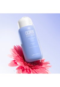 Coco & Eve Boost Therapy volumiserande schampo i en pastellblå flaska med ett vitt lock, placerad ovanför en rosa blomma, med en droppe vätska.