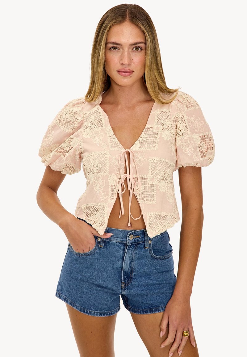 Femme portant un crop top beige à manches courtes bouffantes avec un motif en dentelle, noué sur le devant, et un short en jean bleu taille haute.