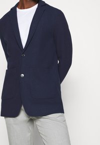Navy blue blazer made of textured fabric, featuring a single button closure, two front pockets, and notched lapels, paired with light pants.
