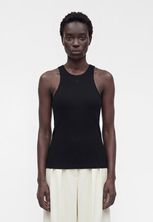 Femme aux cheveux courts et bouclés portant un haut noir sans manches et un pantalon blanc ample, debout devant un fond clair uni.