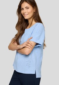 Himmelblaues Texturtop mit kurzen Ärmeln, Rundhalsausschnitt und subtiler Verzierung an der Seite, kombiniert mit dunklen Hosen.