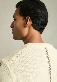 Krems krāsas adīts džemperis ar teksturētu šuvi un brūnu akcentu gar muguras šuvi. Apaļš izgriezums un ribota aproce.