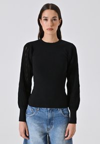 Schwarzer Strickpullover mit langen, strukturierten Puffärmeln. Figurschnitt, runder Ausschnitt, kombiniert mit hellblauen, verwaschenen Jeans.