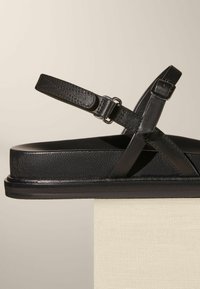 Svart lädersandal med två justerbara korsklingor, platt sula och texturerad yta. Minimalistisk design med en slank silhuett.