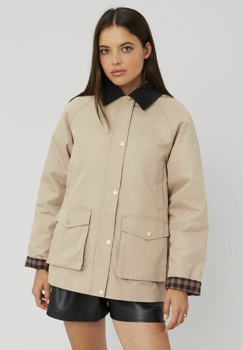 Veste beige avec un col en velours côtelé noir, boutons dorés, deux poches avant et des poignets à carreaux bruns. Texture lisse et design décontracté.