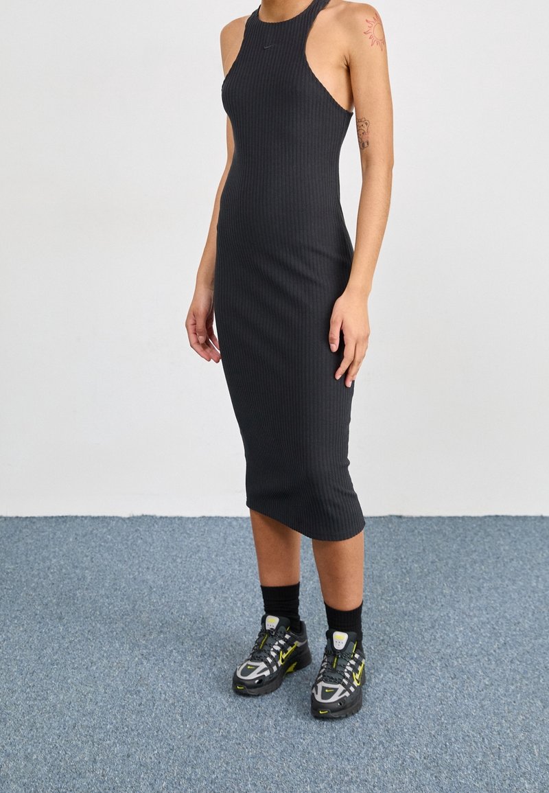 Personne portant une robe midi noire côtelée sans manches avec des baskets noires et jaunes, debout sur un sol recouvert d’un tapis bleu contre un mur blanc.