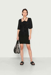 Vestido preto com mangas bufantes e detalhe frontal amarrado, combinado com uma bolsa de rede preta e sandálias deslizantes pretas. Textura de tecido suave.