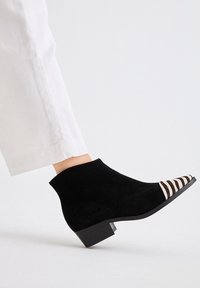 Botín negro de ante hasta el tobillo con punta afilada que presenta un patrón a rayas en blanco y negro. Tacón bajo y cuadrado. Llevado con pantalones blancos.