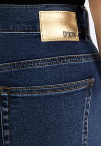Mørkeblå denimjeans med et gyldent lærmerke merket "DRYKORN". Har synlige sømmer langs lommer og livlinje.