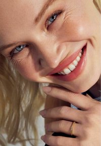 Guldring med små glittrande stenar, bärs på ett finger. Lätt, slät textur; modell som ler, med långt blont hår och klara blå ögon.