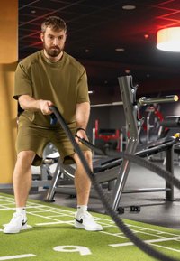 Figura muscolosa in abbigliamento attivo verde oliva che utilizza corde da battaglia nere su un prato verde, con una palestra e macchine per pesi sullo sfondo.