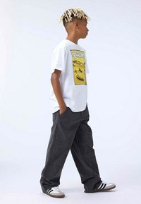 Hvid t-shirt med et gult grafisk design, parret med løstsiddende, mørkegrå cargo bukser og hvide sneakers med sorte striber.