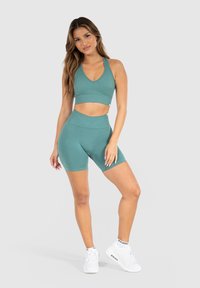 Soutien-gorge de sport en teal avec un décolleté en V et une texture côtelée, associé à un short taille haute assorti. Des baskets blanches complètent la tenue.